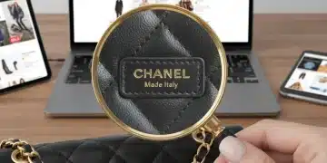 Magnifying glass examining designer label on handbag, laptop with online fashion sites in background, symbolizing online fashion authenticity verification.